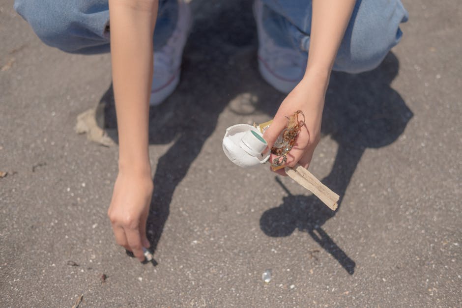 A person crouching on a paved outdoor surface, visible from the waist down, wearing light blue jeans and white sneakers. They are holding a small bundle of trash composed of a white disposable cup with a teal logo, some tangled paper or organic waste, and a wooden stick or stick-like object. The person's right hand is near the ground, while the left hand grips the trash firmly. The background shows the rough gravel of the pavement with no other objects visible. The scene suggests an individual participating in a clean-up activity, highlighting an example of responsible rubbish disposal, which aligns with private waste handling or rubbish removal services like those offered by Rubbish Removal Bromley. The overall setting appears to be an outdoor area, possibly a street or park, where waste is being collected for proper disposal.
