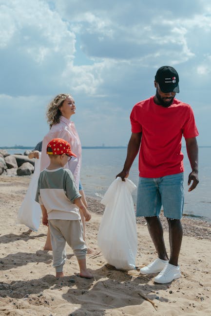 A group of three people on a sandy beach, including a man wearing a red T-shirt, light blue denim shorts, a black baseball cap, and white sneakers, holding a large white plastic bag for rubbish. A young girl with curly blonde hair, dressed in a light pink jacket, stands nearby, looking towards the water, while a boy wearing a red cap, beige trousers, and a grey T-shirt with a white bag over his shoulder watches the man. In the background, there is a calm body of water with rocks lining the shoreline, a partly cloudy sky overhead, and the scene appears to be focused on a clean-up activity, reflecting the importance of private waste disposal services such as those offered by Rubbish Removal Bromley for environmentally responsible rubbish collection.