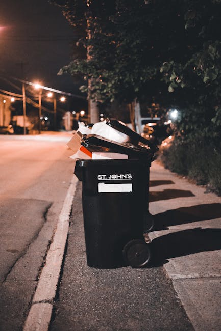 A black plastic wheelie bin with a white label on the front, marked with the name 'St. John's', is positioned on the curbside of a dimly lit street at night. The bin's lid is open and filled with mixed waste, including cardboard boxes and paper, some of which are slightly disorganized and protruding. The surrounding environment features a paved road with a white line marking the edge, and the bin is situated on a concrete pavement adjacent to a grassy verge with dark foliage. In the background, soft orange streetlights illuminate the scene, casting subtle shadows on the pavement and highlighting nearby trees and utility poles. The scene suggests an on-site waste disposal setup, typical of private rubbish collection services, such as those offered by Rubbish Removal Bromley, serving local residents or businesses in the Bromley area. The overall atmosphere is calm, with minimal activity, and the image emphasizes the importance of proper rubbish management and the role of independent waste collection solutions in urban environments.