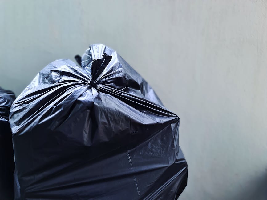 A black plastic wheelie bin with a white label on the front, marked with the name 'St. John's', is positioned on the curbside of a dimly lit street at night. The bin's lid is open and filled with mixed waste, including cardboard boxes and paper, some of which are slightly disorganized and protruding. The surrounding environment features a paved road with a white line marking the edge, and the bin is situated on a concrete pavement adjacent to a grassy verge with dark foliage. In the background, soft orange streetlights illuminate the scene, casting subtle shadows on the pavement and highlighting nearby trees and utility poles. The scene suggests an on-site waste disposal setup, typical of private rubbish collection services, such as those offered by Rubbish Removal Bromley, serving local residents or businesses in the Bromley area. The overall atmosphere is calm, with minimal activity, and the image emphasizes the importance of proper rubbish management and the role of independent waste collection solutions in urban environments.