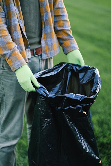 A person standing outdoors on a grassy area, holding open a large black plastic rubbish bag with both hands, wearing light green gloves and casual clothing including a plaid shirt with yellow, grey, and blue tones and grey trousers. The background features a well-maintained green lawn, suggesting a garden or park setting. The scene is well-lit with natural daylight. This image reflects the process of waste collection, where an individual prepares rubbish for disposal or collection, supporting the context of private waste handling and alternative rubbish removal services provided by companies like Rubbish Removal Bromley. The focus on the rubbish bag and gloves emphasizes cleanliness and proper waste management practices outside of official municipal services.