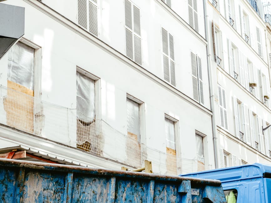The image shows a large, open-top metal waste skip with a weathered, rusty surface painted in a faded blue color, situated on a street or alleyway in front of a building. The skip contains various types of rubbish including cardboard boxes, plastic bags, and loose debris, with some items partially spilling over the edges. Behind the skip, part of a blue commercial vehicle is visible, likely used for rubbish collection or waste disposal. In the background, there is a multi-storey residential building with white-painted walls and multiple window openings, some of which are boarded up or fitted with protective netting. The windows feature closed, louvered shutters, and small planters can be seen on some window sills, indicating the building’s residential use. The environment appears to be urban, with natural daylight illuminating the scene, highlighting the textures of the materials and the slightly grimy or aged appearance of the building façade. The setting subtly implies an alternative waste handling scenario, with Rubbish Removal Bromley potentially providing private rubbish collection services that differ from statutory local authority disposal methods.