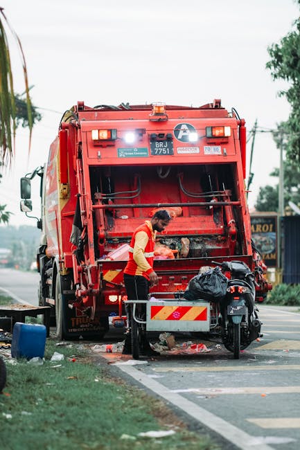 A sanitation worker wearing a yellow and red uniform is positioned behind a large, red waste collection vehicle with its rear compartment open on a roadside. The worker appears to be loading or sorting rubbish, with a black trash bag placed on a small platform attached to the vehicle. The vehicle has visible warning and identification markings, including the code 'BRJ 7751' and reflective yellow and red stripes on the rear. The scene is set on a street with a curb and road markings, with a grassy verge to the left and some scattered debris near the base of the vehicle. In the background, there are trees, utility poles, and a building with signage, suggesting an urban or suburban environment. The lighting indicates a bright, overcast day, highlighting the details of the waste collection process, which is an example of independent waste handling and rubbish removal typically managed by private service providers like Rubbish Removal Bromley.