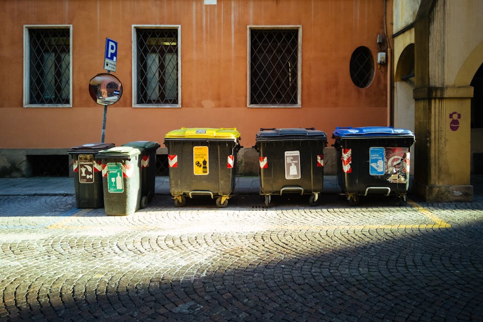 Four wheeled rubbish bins are positioned on a cobblestone pavement in front of a peach-colored building with three small windows protected by metal grilles. The first bin on the left is green with a black lid and features a white icon of a bottle and a recycling symbol. The second bin has a yellow lid with yellow and black labels indicating general waste with a biohazard symbol. The third bin has a black lid with a label depicting a glass bottle and a trash can icon. The fourth bin has a blue lid with stickers showing a recycling symbol, a hand placing waste into a container, and a graphic of a lunchbox or containers. A convex mirror is mounted on a pole next to the first bin, reflecting part of the street. The scene is lit by natural light, casting shadows across the cobblestones, suggesting daytime. The background includes a worn wall, part of a building with an archway on the right and a small round black window. The image is related to waste management services, illustrating a typical outdoor waste disposal setup, as managed by local rubbish removal providers like Rubbish Removal Bromley, showcasing different waste segregation containers for different materials.