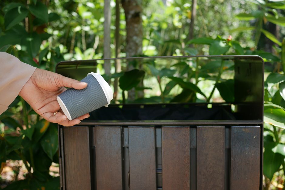 A person wearing a beige long sleeve shirt is holding a grey, ridged paper coffee cup with a white lid, about to place it into a large, open, black metal outdoor rubbish bin. The bin is situated on a paved surface within a garden or green outdoor area, with green foliage, tall plants, and tree trunks visible in the background. The interior of the bin appears empty or lightly filled, and the setting suggests an environment related to outdoor waste disposal or rubbish collection, possibly indicating an alternative waste handling method outside standard council services. The scene is well-lit with natural daylight, emphasizing the contrast between the dark metal bin and the vibrant greenery surrounding it.